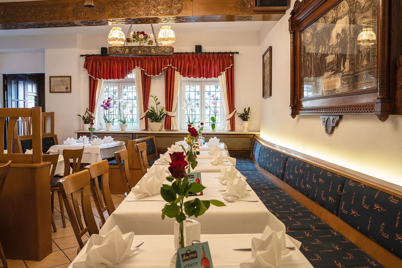 Lichtdurchfluteter Gastraum mit traditionellen Details und gemütlicher Atmosphäre Heller Restaurantbereich mit roten Vorhängen, Blumen und langen Tischen